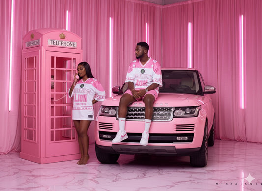 Two people in matching pink outfits sitting on a pink Range Rover with a pink telephone booth in the background.