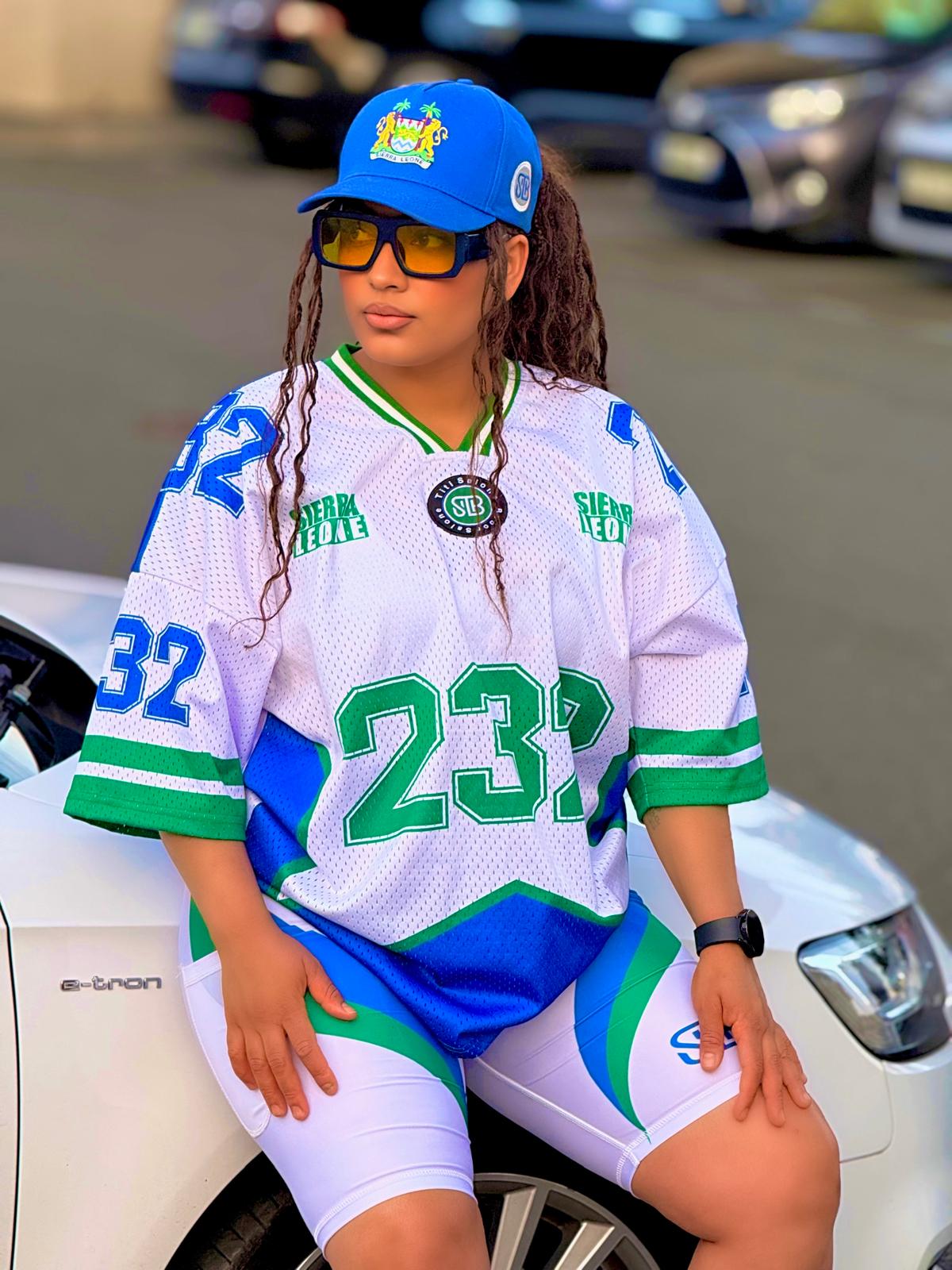 Person wearing a sports jersey and cap sitting on a car in a parking lot.