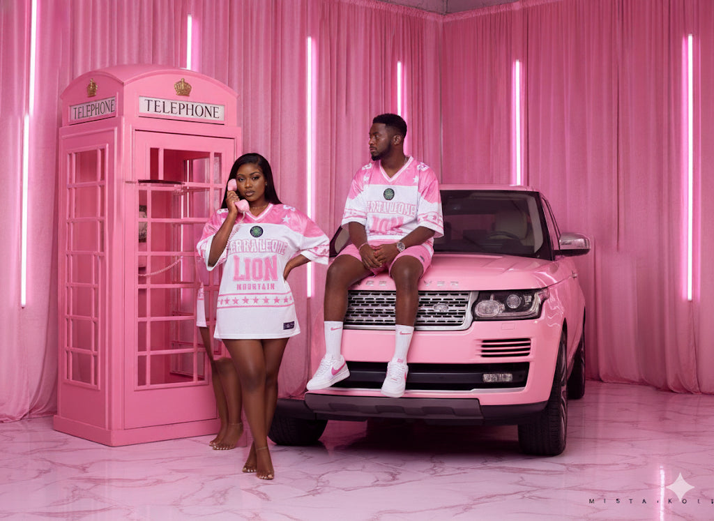 Two people in matching pink outfits sitting on a pink car with a pink telephone booth in the background.