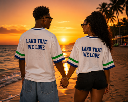 A couple holding hands from behind on a beach at sunset, wearing matching white t-shirts with blue and green striped sleeves and the text 'LAND THAT WE LOVE' printed in blue on the back.