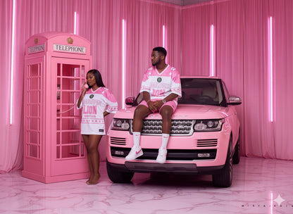 Two people in matching pink outfits sitting on a pink Range Rover with a pink telephone booth in the background.