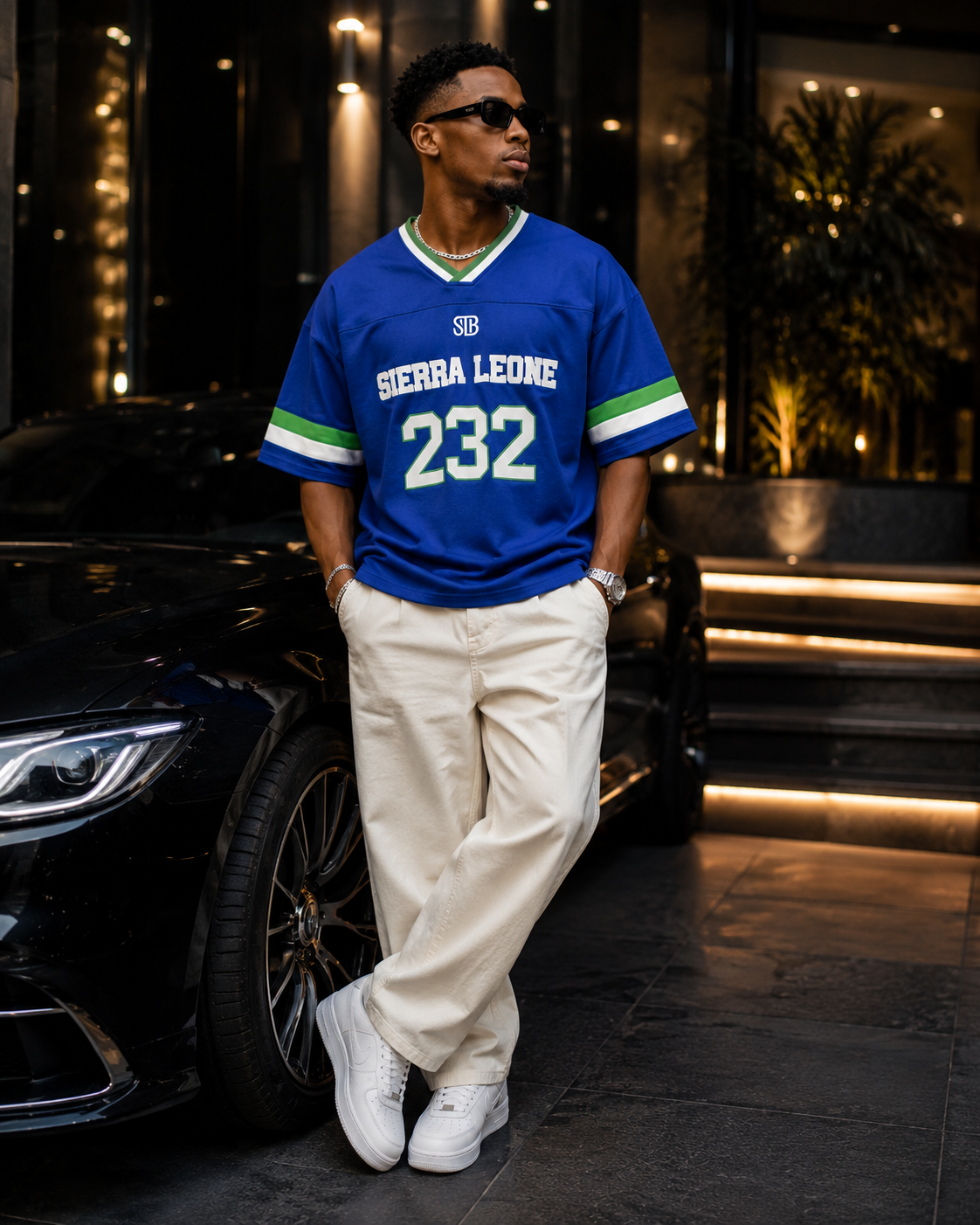 A man wearing a blue and green Sierra Leone soccer jersey with the number 232, cream trousers, and white sneakers, standing next to a black car at night.