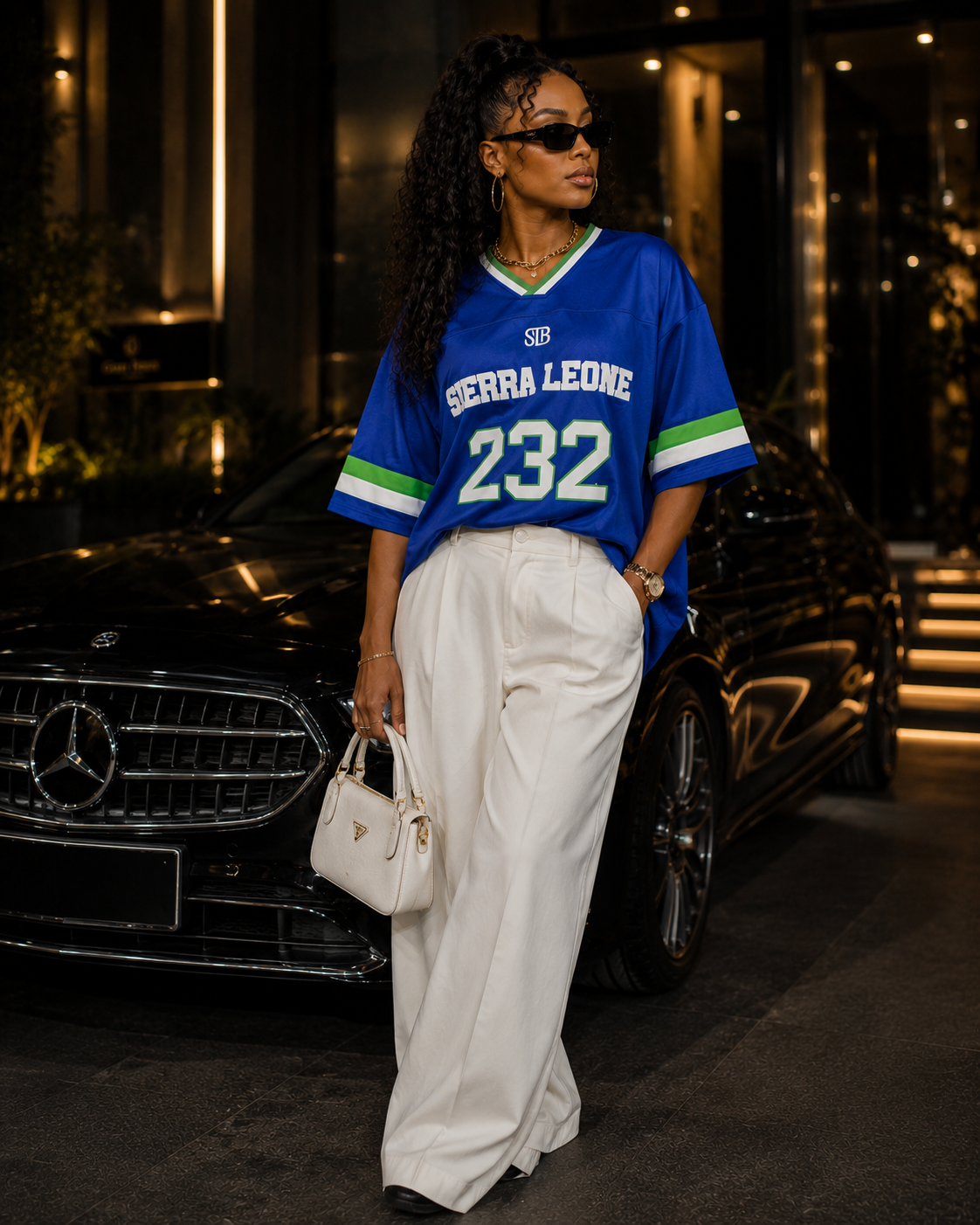 A woman stands in front of a black Mercedes-Benz car at night, wearing an oversized blue sports jersey with 'SIERRA LEONE 232' on the front, paired with white wide-leg trousers and carrying a small white handbag.