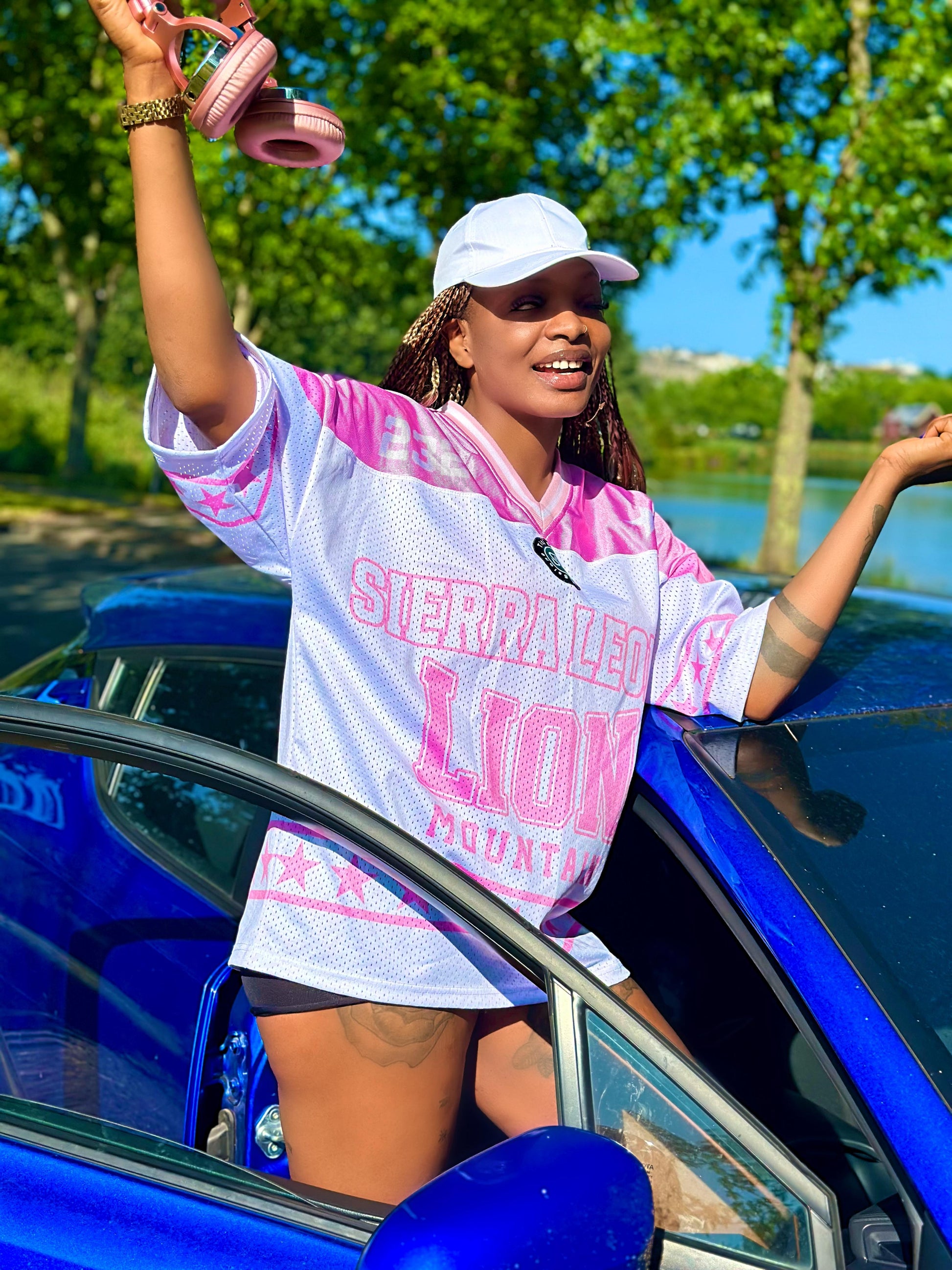 Woman in a tie-dye shirt and cap standing next to a blue car with trees and water in the background