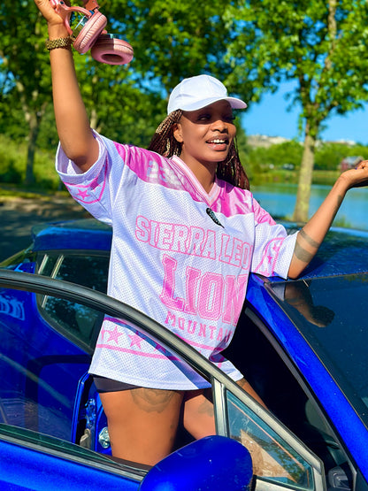Woman in a tie-dye shirt and cap standing next to a blue car with trees and water in the background