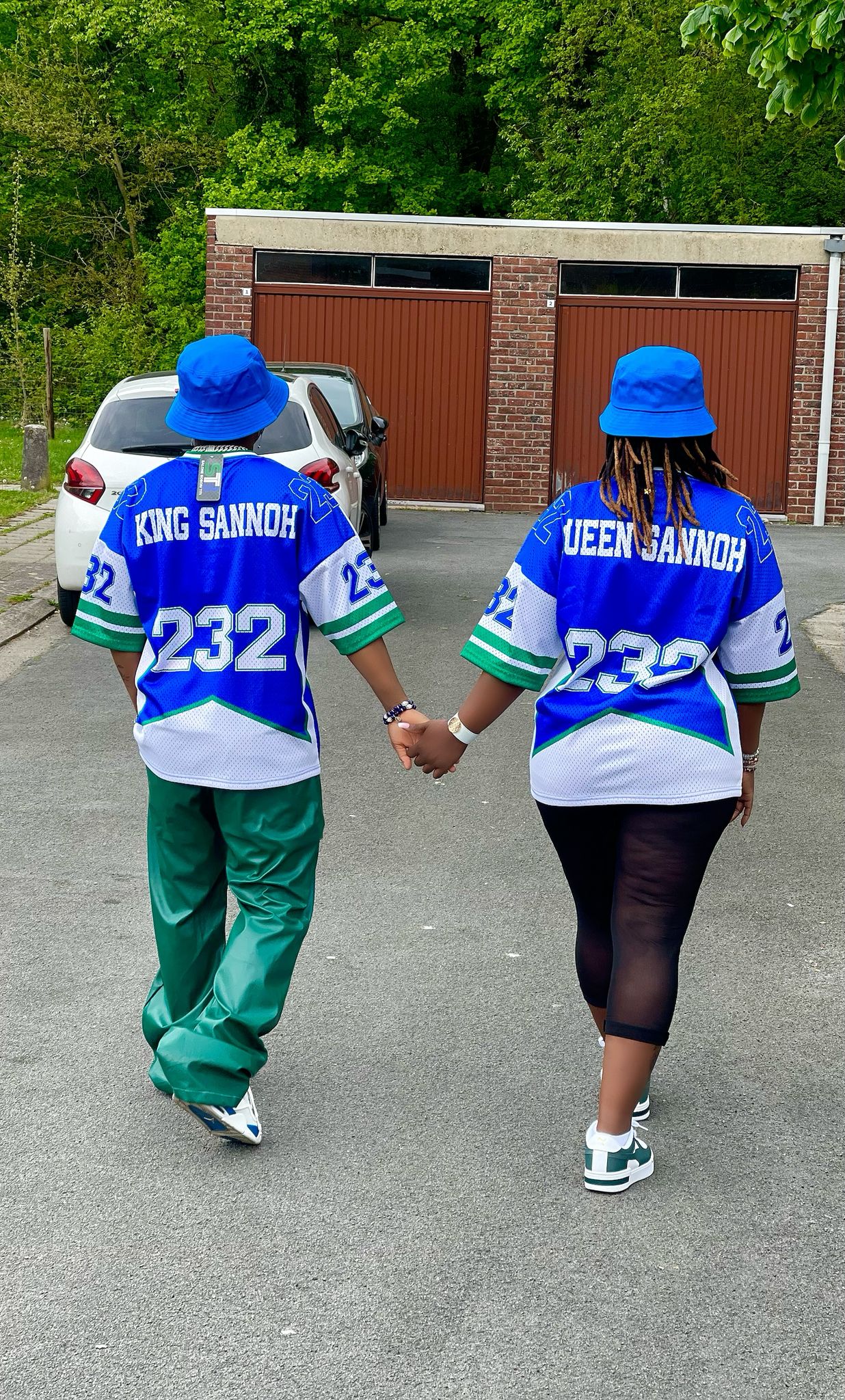 Two people wearing matching blue and green outfits with 'King Sannoh' and 'Queen Sannoh' text, walking hand in hand on a street.