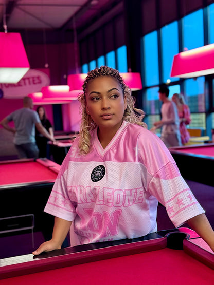 Woman in a pink jersey standing behind a pool table in a pool hall.